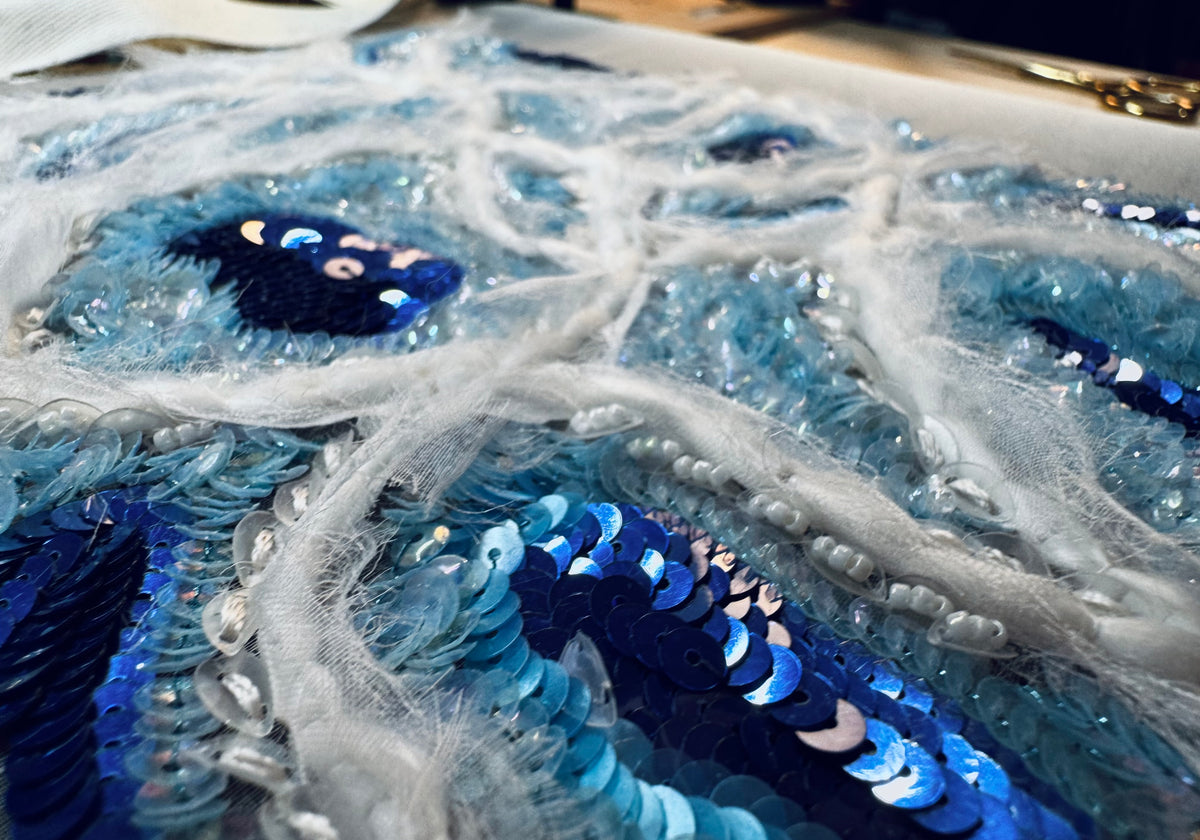 Close-up of a heavily embellished blue sequined textile or garment surface with layered tulle, bead strands, and reflective sequins in navy, cobalt, and light blue.