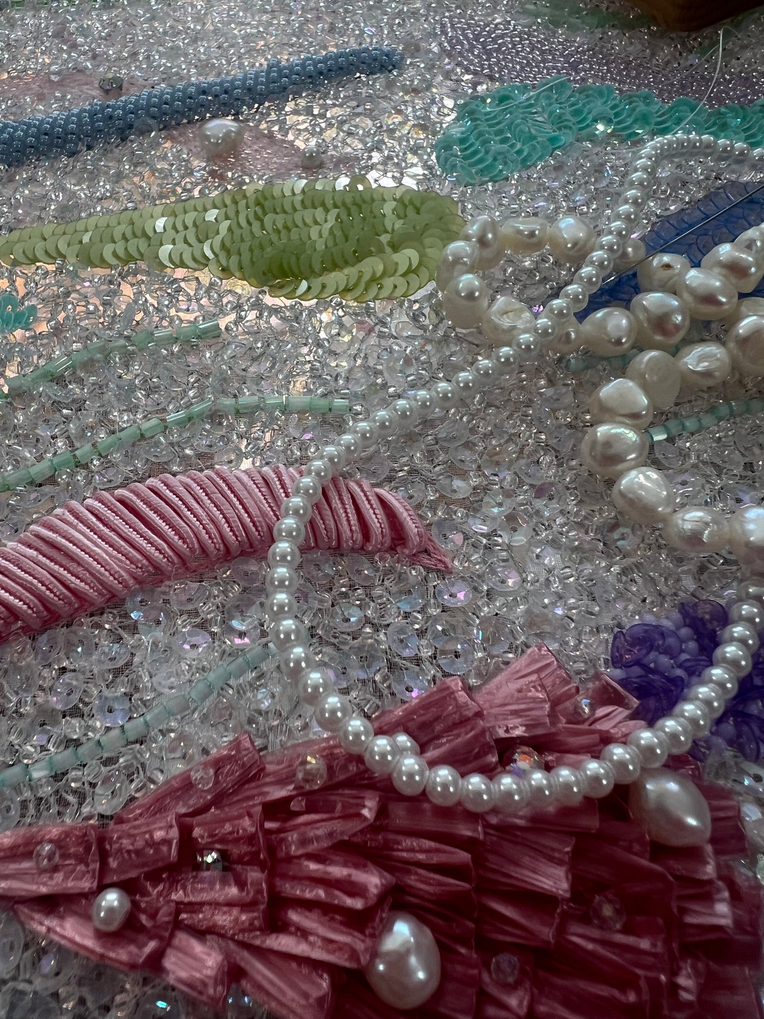A close-up of a handmade beaded and sequined textile piece featuring white pearl beads, pink and green sequins, and silver rhinestones on a glittery background.